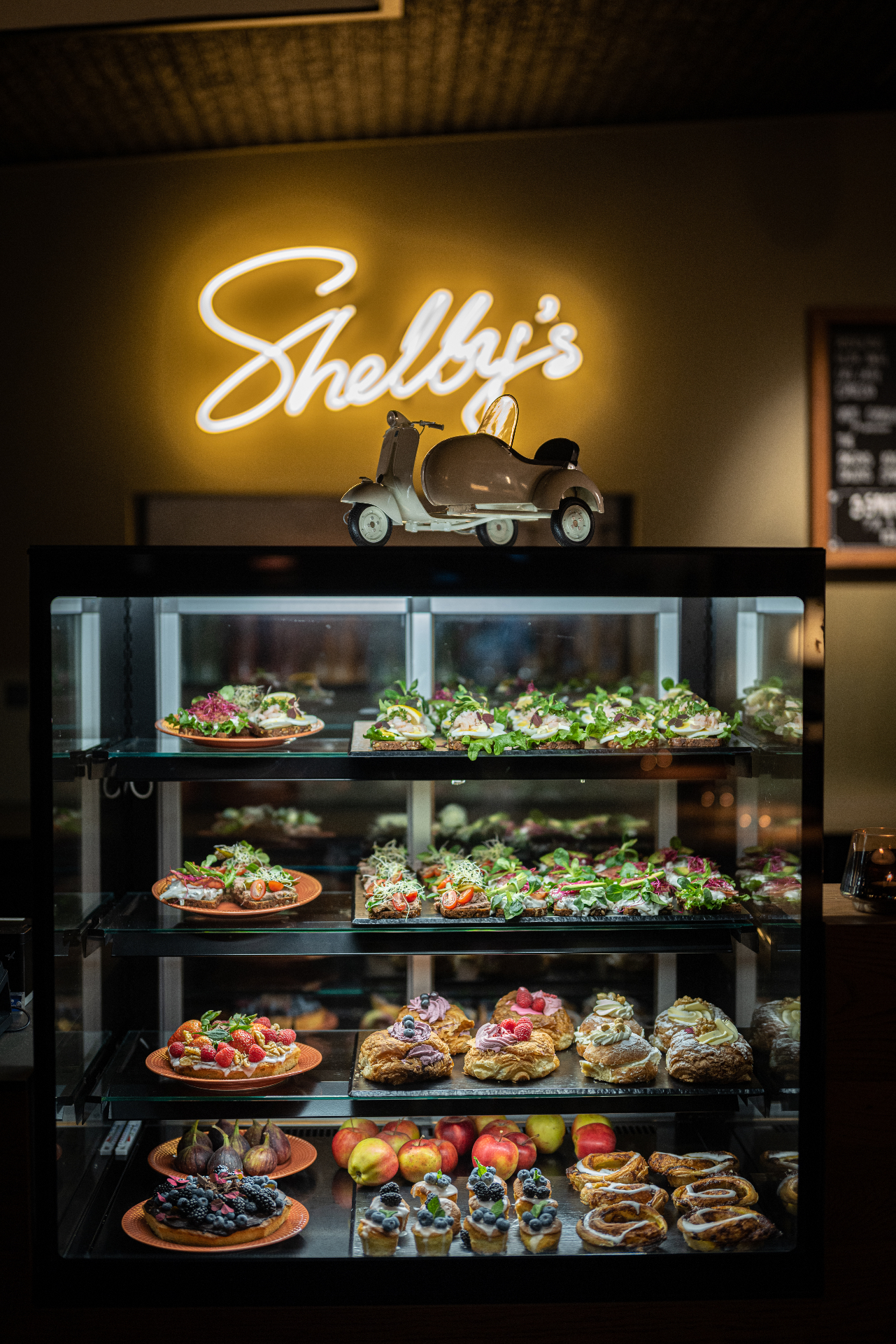 Shelby's Café — kagedisk med smørrebrød, wienerbrød og frugttærter under neonskilt